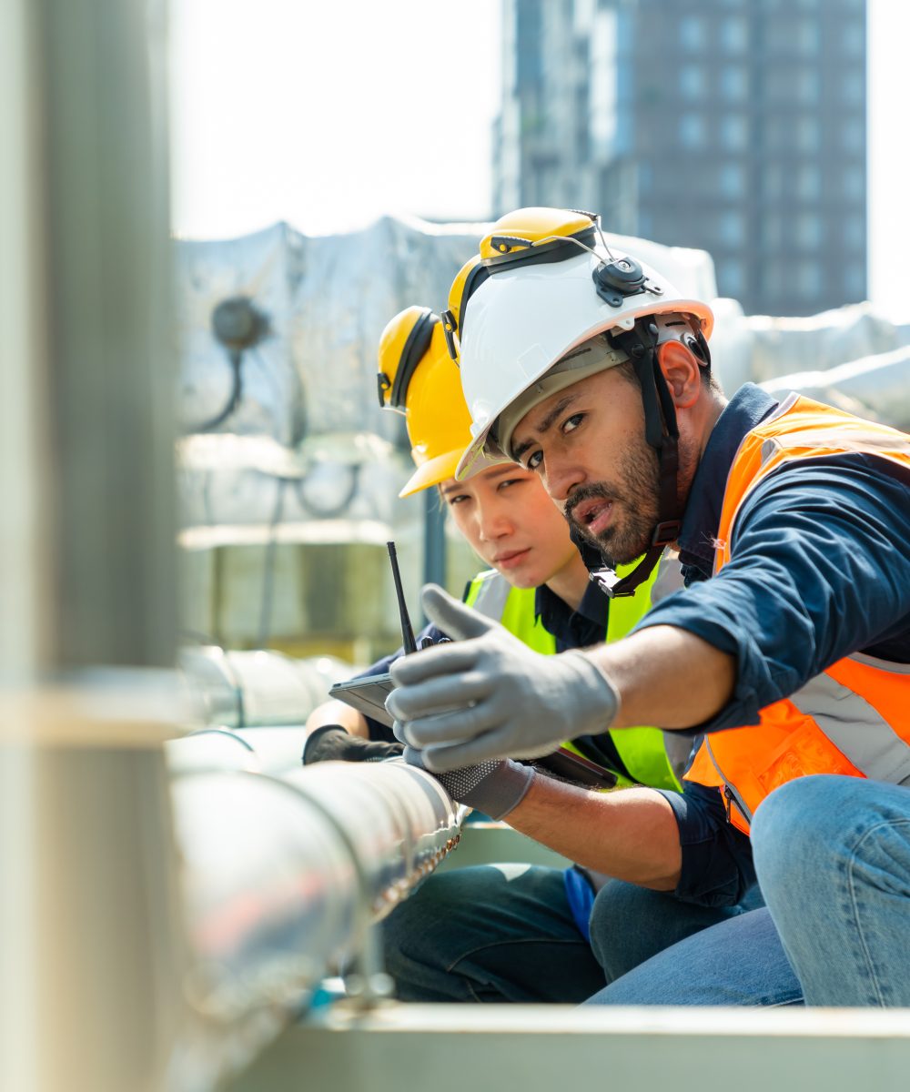 Professional Asian man and woman teamwork engineer in safety uniform working at outdoor construction site rooftop. Industrial technician worker maintenance checking building exterior plumbing systems.