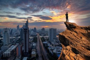 Concept vision, Businessman wearing comfortable casual suit jacket standing chest hug on top of peak mountain and looking forwards with cityscape background, success, competition and leader concept.